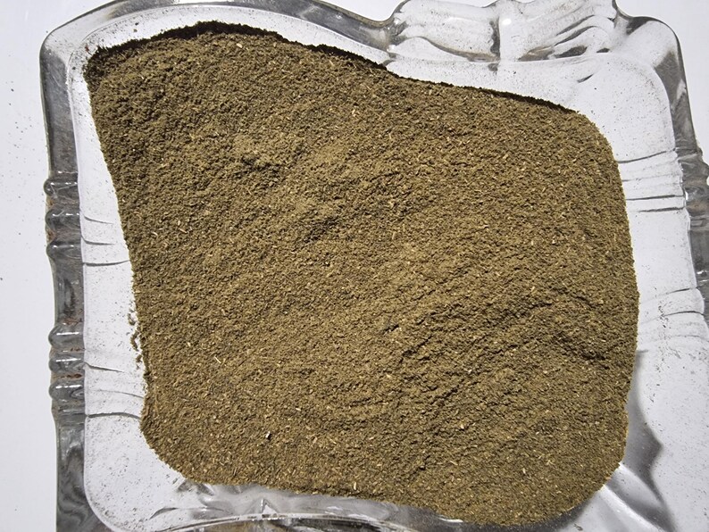 May include: A finely ground, earthy brown herbal powder contained within a clear, square glass dish with decorative scalloped edges. The texture of the powder appears uniform and dry, filling the center of the transparent glass container.
