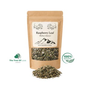 Op de afbeelding: Een bruine kraftpapieren zak met gedroogd frambozenblad, voorzien van de tekst Raspberry Leaf (Rubus idaeus) en een berggrafiek. Een kleine hoop van de groene, gefragmenteerde kruidenbladeren ligt voor de zak.