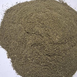 May include: A mound of finely ground, earthy-toned herbal powder with a coarse, granular texture. The substance displays a mix of muted olive green and brownish particles, appearing as a dry, loose botanical material piled into a rounded heap.