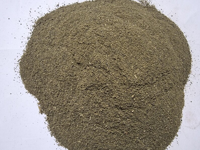 May include: A mound of finely ground, earthy-toned herbal powder with a coarse, granular texture. The substance displays a mix of muted olive green and brownish particles, appearing as a dry, loose botanical material piled into a rounded heap.