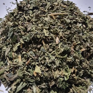 May include: A pile of dried, crushed green leaves and small woody stems. The texture is coarse and fragmented, featuring various shades of dark forest green, olive, and brown. The plant matter is scattered in an irregular, mounded shape.