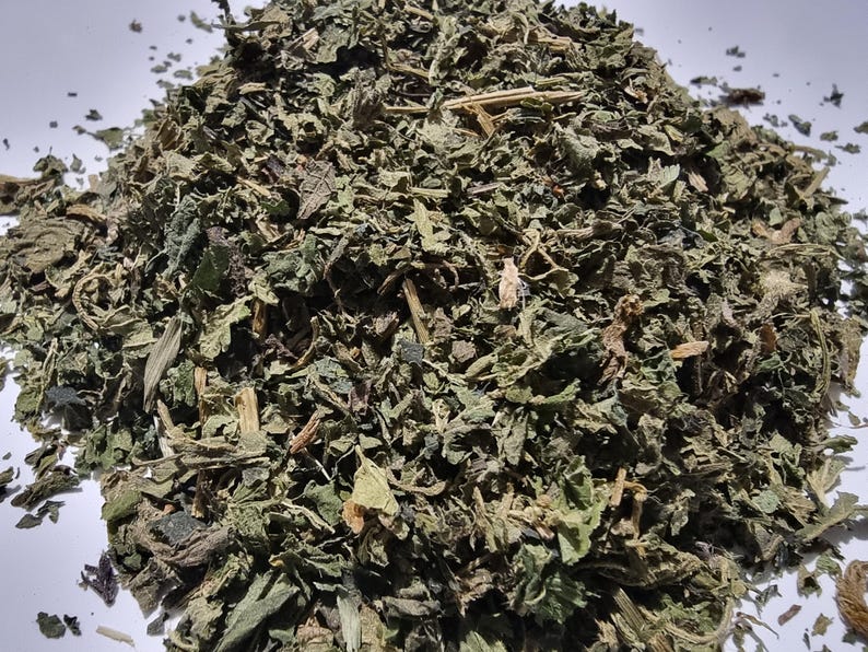 May include: A pile of dried, crushed green leaves and small woody stems. The texture is coarse and fragmented, featuring various shades of dark forest green, olive, and brown. The plant matter is scattered in an irregular, mounded shape.