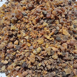 May include: A collection of small, irregular resin pieces in various shades of amber, brown, and dark gray. The texture is crystalline and granular, with some translucent golden fragments mixed among the darker, opaque, and earthy-toned particles.