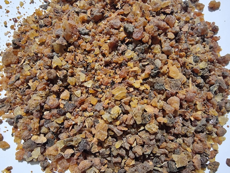 May include: A collection of small, irregular resin pieces in various shades of amber, brown, and dark gray. The texture is crystalline and granular, with some translucent golden fragments mixed among the darker, opaque, and earthy-toned particles.