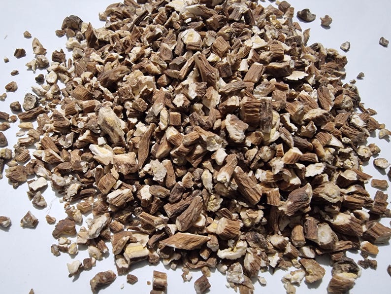 May include: A pile of dried, chopped root pieces featuring a mix of dark brown bark and light cream-colored interiors. The fragments are irregular in shape and size, scattered across a plain white surface, showing a fibrous and woody texture.