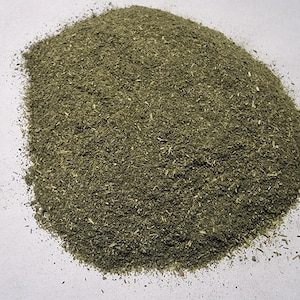 May include: A mound of finely ground, dried green herbal powder with a coarse, textured consistency. The mixture displays various shades of olive and forest green, featuring small, visible flecks of dried plant matter throughout the pile.