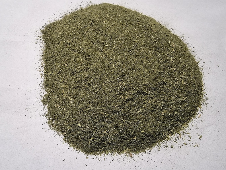 May include: A mound of finely ground, dried green herbal powder with a coarse, textured consistency. The mixture displays various shades of olive and forest green, featuring small, visible flecks of dried plant matter throughout the pile.