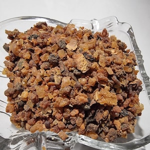 May include: A clear glass bowl filled with a collection of small, irregular, resinous chunks in shades of amber, brown, and dark reddish-brown. The pieces have a crystalline, translucent, and matte texture, varying in size and shape throughout.