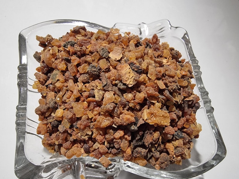 May include: A clear glass bowl filled with a collection of small, irregular, resinous chunks in shades of amber, brown, and dark reddish-brown. The pieces have a crystalline, translucent, and matte texture, varying in size and shape throughout.