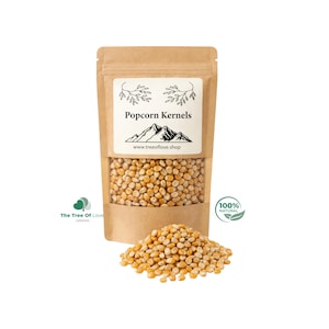 Puede incluir: Una bolsa de papel kraft con granos de palomitas de maíz, con una ventana transparente, una etiqueta blanca con el texto "Popcorn Kernels" y una ilustración de montaña. Un pequeño montón de granos está delante de la bolsa.