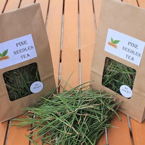 Può includere: Due sacchetti di carta marrone contenenti tè di aghi di pino, ciascuno con un'etichetta bianca con la scritta PINE NEEDLES TEA e un logo biologico. Un sacchetto indica 85 g e l'altro 114 g. Un mucchio di aghi di pino verdi sfusi si trova al centro.