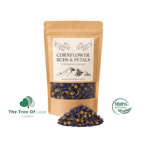 Op de afbeelding: Een bruine kraftpapieren zak met gedroogde korenbloemknoppen en bloemblaadjes. Het etiket vermeldt CORNFLOWER BUDS & PETALS (Centaurea cyanus) met een bergafbeelding en websiteadres. Een hoopje blauwe en bruine gedroogde bloemdelen ligt ervoor.