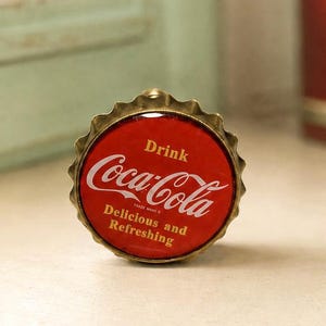 May include: A circular metal bottle cap with a red background and a gold-toned scalloped edge. The text "Drink Coca-Cola" is printed in white script, with the words "Delicious and Refreshing" in smaller white lettering underneath the logo.