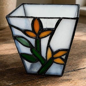 May include: A square stained glass planter featuring a white background with a floral motif. The design includes orange petals and green leaves outlined in black solder. The vessel has a tapered shape with four flat sides and a wide open top.