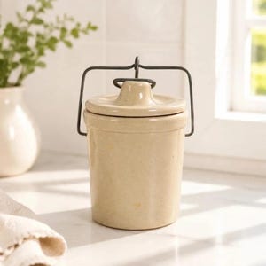 May include: A cylindrical cream-colored ceramic crock with a matching lid and a dark metal wire bail handle. The container features a smooth, solid-colored finish and a central knob on the lid, providing a simple and functional storage vessel.