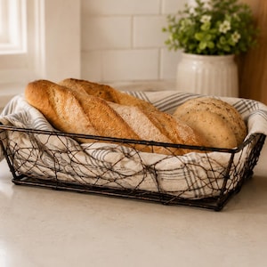 May include: A rectangular dark brown wire basket lined with a white and gray striped fabric cloth. Inside the basket are two golden-brown crusty baguettes and a round loaf of bread, all resting on the fabric liner within the metal wire frame.