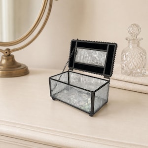 May include: A rectangular glass jewelry box with a black metal frame and textured, semi-opaque glass panels. The lid is hinged with a delicate metal chain and features a central rectangular glass insert. The box stands on four small, rounded feet.