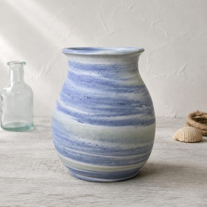 May include: A ceramic vase with a rounded body and a flared rim, featuring a marbled pattern of horizontal blue, white, and light gray streaks. The surface has a matte finish with subtle dark speckles distributed throughout the blue and white bands.