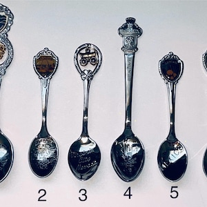 May include: Six vintage souvenir spoons with decorative handles. Each spoon has a different design on the handle, including shields, a covered wagon, and a crown. The spoon bowls are engraved with city names.