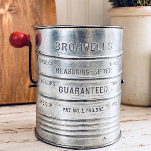 May include: A cylindrical metal flour sifter with a red wooden crank handle. The body features embossed text reading BROMWELL'S MEASURING-SIFTER, MADE IN U.S.A., GUARANTEED, and PAT. NO. 1.753,995, along with measurement markings for one, two, and three cups.