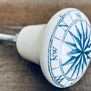 May include: White ceramic cabinet knob with a blue compass rose design. The knob has a rounded shape and a metal screw for attachment. The compass rose features directional markings and a starburst pattern.