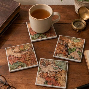 May include: A set of four square coasters featuring a vintage map design with muted tones of orange, green, and beige. Each coaster has a smooth surface and white edges. One coaster supports a beige ceramic mug filled with a brown liquid beverage.