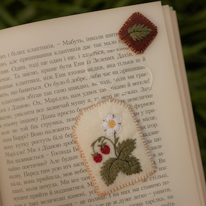 May include: Two handmade felt bookmarks rest on an open book. One is a brown square with a green leaf design. The other is a cream-colored bookmark with a strawberry plant design, featuring a white flower and red berries.