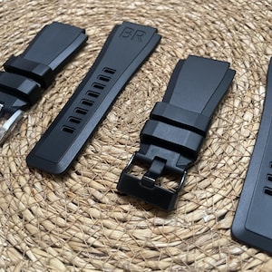 May include: Four black watch straps with silver buckles and the letters "BR" embossed on the straps. The straps are made of a flexible material and have a textured surface. The straps are displayed on a woven, tan surface.