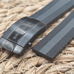 Könnte beinhalten: Nahaufnahme eines grauen Uhrenarmbands mit schwarzer Schließe. Auf der Schließe steht "BREITLING". Das Armband hat eine strukturierte Oberfläche mit einem grauen Mittelstreifen.