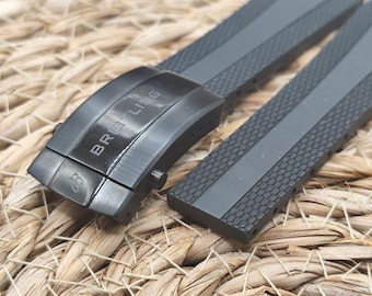 May include: A close-up of a gray watch strap with a black clasp. The clasp is engraved with the word "BREITLING". The strap has a textured surface with a central gray stripe.