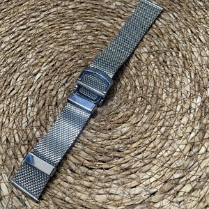 May include: A silver stainless steel watch strap with a mesh design. The strap has a secure clasp and is shown against a woven, textured background. The strap is designed to be durable and stylish.