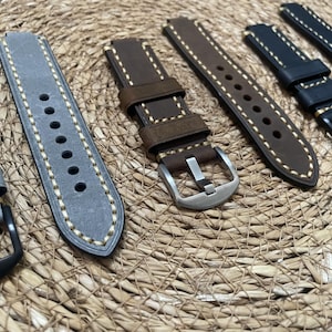 May include: A selection of watch straps in different colours and materials. The straps include grey suede, brown leather, and black leather, all with contrasting stitching. Buckles are silver and black.