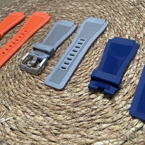 May include: A selection of watch straps in shades of orange, grey, and blue. The straps are made from a flexible material and have adjustment holes and metal buckles. The straps are presented on a woven surface.