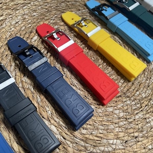 Könnte beinhalten: Eine Auswahl an farbenfrohen Uhrenarmbändern in Schwarz, Marineblau, Rot, Gelb und Blau. Jedes Armband hat eine Metallschnalle und den Markennamen auf dem Gummi geprägt. Die Armbänder sind auf einer gewebten Oberfläche angeordnet.