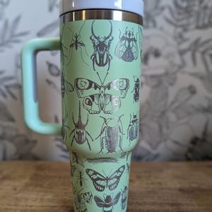 May include: A mint green travel mug with a handle, lid, and a silver rim. The mug is decorated with black illustrations of various insects, including butterflies, beetles, and ants. The handle and lid button are also mint green.