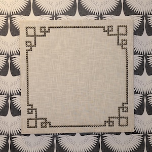 May include: A square, off-white linen placemat with a decorative border of small, dark brown studs. The placemat is set against a wallpaper background featuring a repeating pattern of white birds on a dark gray background.