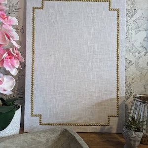 May include: Rectangular white linen wall art with a gold studded border. The gold studs create a decorative frame around the linen. The wall art is displayed on a wooden surface.