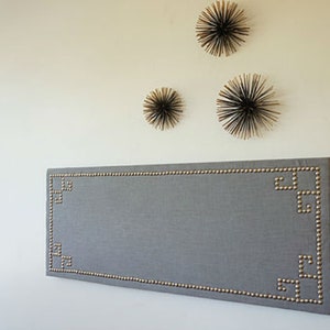 May include: A rectangular gray headboard with brass-colored nailhead trim. The headboard has a geometric design in the corners. Above the headboard are three decorative starburst wall hangings.
