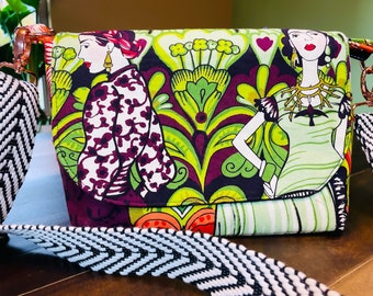 May include: A rectangular crossbody bag with a colorful print featuring stylized figures and floral patterns in green, purple, and white. The bag has a black and white chevron strap and rose gold hardware.