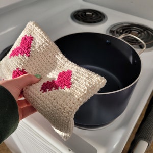 May include: A white crocheted pot holder with pink hearts. The pot holder is being held over a black pot on a white stovetop.