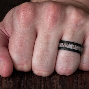 May include: A black ring with a hammered finish and a central inlay featuring a textured, light brown and grey organic material. The band has a flat profile and a wide design, showcasing a contrast between the dark metal and the natural inlay.