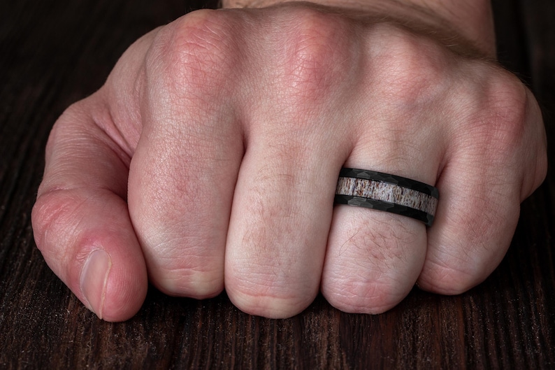 May include: A black ring with a hammered finish and a central inlay featuring a textured, light brown and grey organic material. The band has a flat profile and a wide design, showcasing a contrast between the dark metal and the natural inlay.