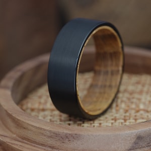 May include: A black tungsten carbide ring with a wooden inlay. The ring is resting on a wooden surface.