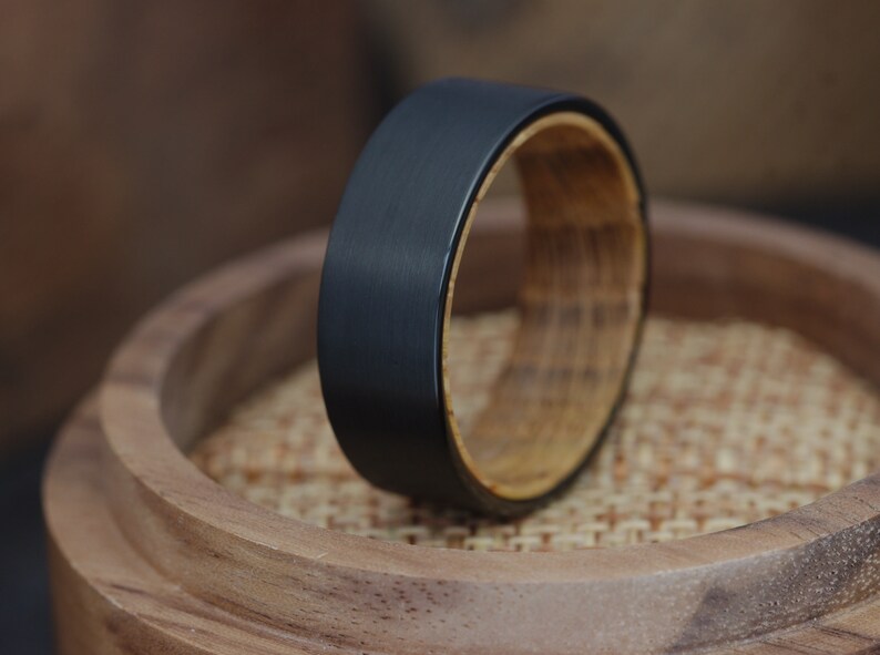 May include: A black tungsten carbide ring with a wooden inlay. The ring is resting on a wooden surface.