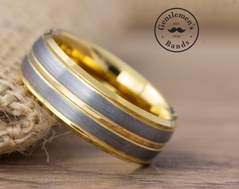 May include: A gold and grey tungsten carbide ring with two gold bands. The ring is on a brown surface.