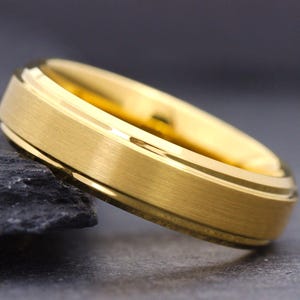 May include: A gold-colored ring with a brushed matte finish on the outer band and polished, shiny beveled edges. The ring features a circular design with a smooth interior surface and a distinct metallic luster, showcasing a clean and simple band.
