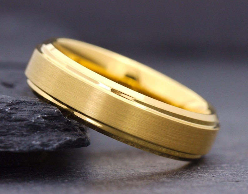 May include: A gold-colored ring with a brushed matte finish on the outer band and polished, shiny beveled edges. The ring features a circular design with a smooth interior surface and a distinct metallic luster, showcasing a clean and simple band.