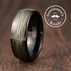 Op de afbeelding: Een zwarte ring met een getextureerd, lichtgekleurd patroon. De ring heeft een glanzende afwerking en staat tegen een houten achtergrond. De ring is van Gentlemen's Bands, Est. 2019.