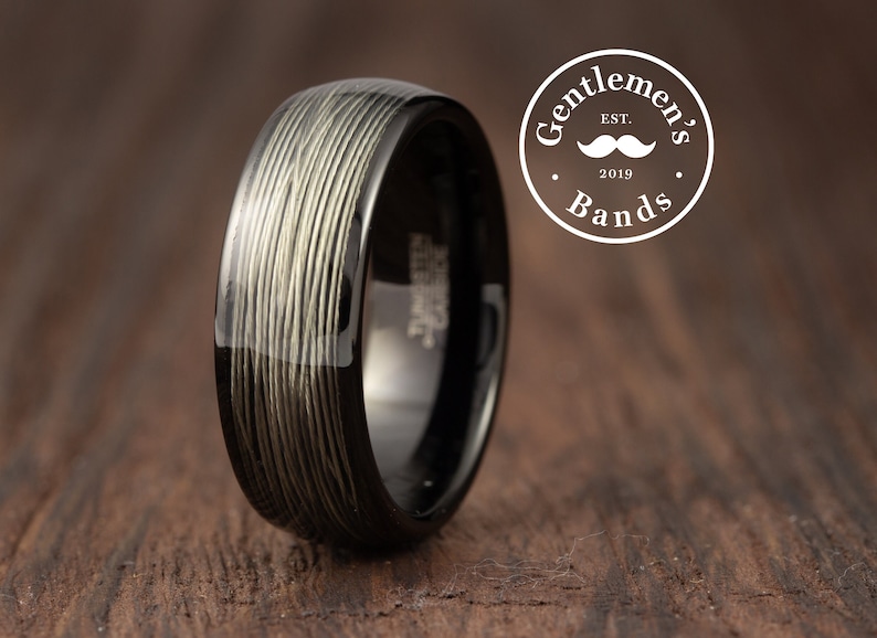 Op de afbeelding: Een zwarte ring met een getextureerd, lichtgekleurd patroon. De ring heeft een glanzende afwerking en staat tegen een houten achtergrond. De ring is van Gentlemen's Bands, Est. 2019.