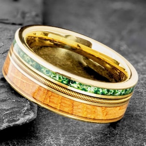 Puede incluir: Un anillo dorado con un diseño único. El anillo presenta una banda de madera marrón claro, una incrustación de piedra verde y un fino alambre de metal enrollado. El anillo está sobre un fondo oscuro y texturizado.